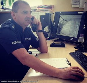 Policjant w mundurze, siedzi przy biurku na którym stoją ustawione monitory komputera, policjant trzyma przy uchu słuchawkę do telefonu