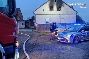 Policyjny radiowóz oraz wóz strażacki na tle budynku w którym ugaszono pożar. Widzimy policjanta i strażaka oraz osobę cywilną