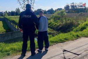umundurowany policjant obok sprawca rozboju z rękami do przodu . W oddali widać zabudowania i łąki