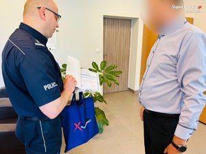 zdjęcie ze spotkania - policjant z mężczyzną w pokoju biurowym, policjant wręcza mężczyźnie dyplom i upominek w niebieskiej torbie
