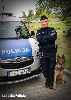 Umundurowany policjant stojący z psem służbowym przy radiowozie policyjnym
