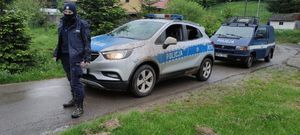 umundurowany policjant, za nim dwa radiowozu. W tle drzewa i domy