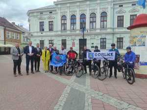 Rowerzyści oraz kilka innych osób stoi trzymając flagi IPA na tle budynku Urzędu Miasta w Sanoku