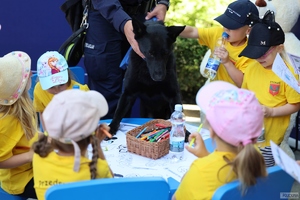 grupka dzieci przy stoliku, na którym znajdują się kartki papieru i kolorowe kredki, obok stolika pies z policjantem