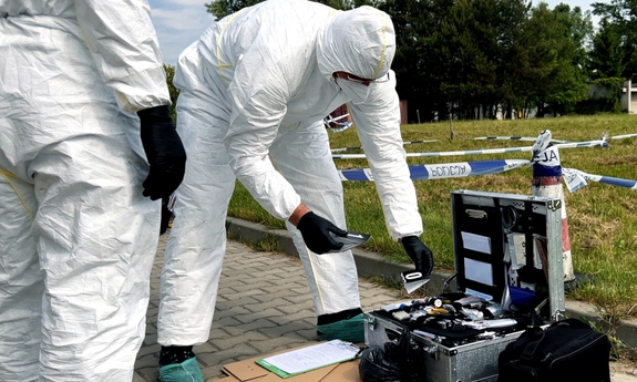 dwaj technicy kryminalistyki w białych kombinezonach, obok leży otwarta metalowa walizka ze sprzętem