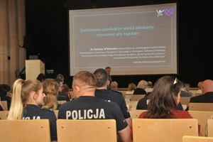 Zdjęcie pleców siedzących w rzędach krzeseł policjantów.