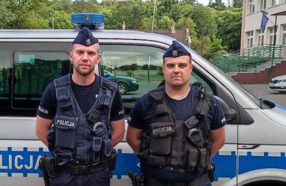 dwóch umundurowanych policjantów stoi przy radiowozie