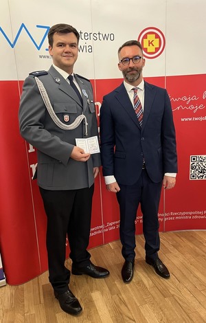 umundurowany policjant pozuje do zdjęcia trzymając legitymację honorowego dawcy krwi zasłużonego da zdrowia narodu. Obok niego stoi mężczyzna w garniturze