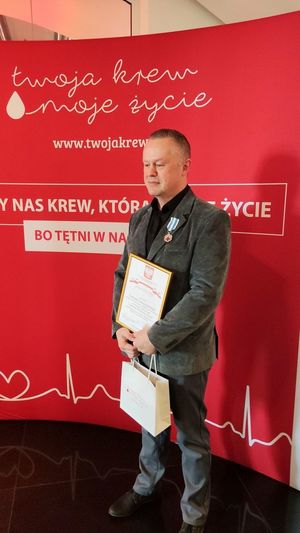 policjant stoi na tle baneru, w klapie marynarki ma przypiętą odznakę, w rękach trzyma list gratulacyjny w ramkach i papierową torbę z upominkami