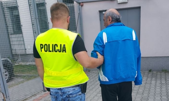 policjant w żółtej kamizelce z napisem policja prowadzi zatrzymanego mężczyznę w niebieskiej bluzie