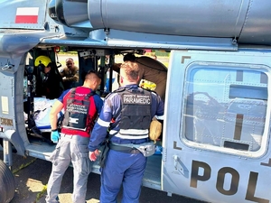 Dwaj ratownicy stoją przy śmigłowcu. Pacjent leży w jego wnętrzu, W głębi ratownik przy noszach pacjenta i policyjni lotnicy