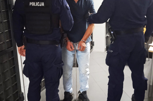 Dwóch umundurowanych policjantów prowadzi zatrzymanego mężczyznę, który ma założone kajdanki zespolone na ręce i nogi trzymane z przodu