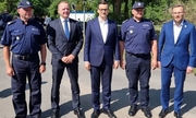Premier RP Mateusz Morawiecki, dwaj policjanci i dwaj inni mężczyźni stojący na drodze. W tle drzewa.