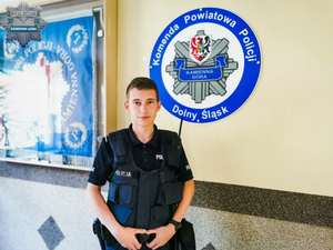 Policjant stoi na korytarzu, na ścianie w tle logo i napis Komenda Powiatowa Policji Kamienna Góra