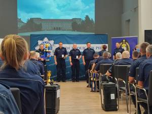 policjanci uczestniczący w uroczystym zakończeniu konkursu. Na sali siedzą policjanci na krzesłach. Przed nimi stoją w szeregu policjanci, którym wręczane są wyróżnienia