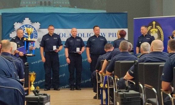 policjanci uczestniczący w uroczystym zakończeniu konkursu. Na sali siedzą policjanci na krzesłach. Przed nimi stoją w szeregu policjanci, którym wręczane są wyróżnienia