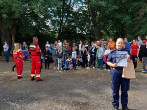 Policjantka trzyma w rękach plakat akcji &quot;Kręci mnie bezpieczeństwo nad wodą&quot;, w tle ratownicy medyczni rozmawiają z grupą dzieci.