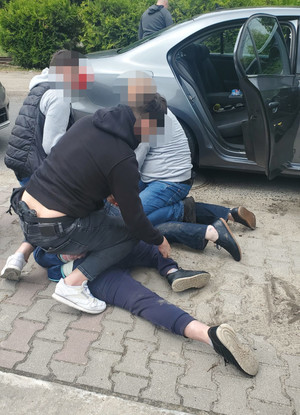 trzej policjanci w trakcie zatrzymywania mężczyzny, który leży na ziemi, obok stoi samochód