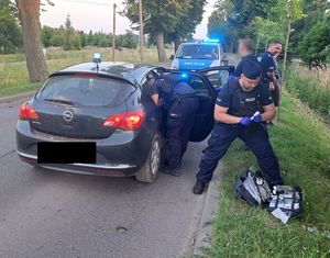 policyjny radiowóz i auto osobowe stoją na jezdni. Przy aucie widać trzech policjantów, z których jeden stoi na poboczu z ubraną po cywilnemu osobą, drugi trzyma opatrunek a przed nim na trawie leży apteczka, a trzeci nachyla się nad siedzeniem pasażera z tyłu.