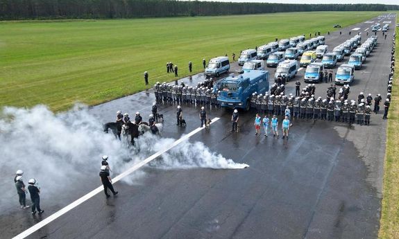 widok z drona na płytę lotniska: na płycie widoczni rozstawieni policjanci z oddziałów prewencji, widoczny jest wóz bojowy oraz policyjne radiowozy, policjanci z zespołu antykonfliktowego, policjanci na koniach i z psami