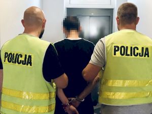 Odwróceni tyłem dwaj policjanci w żółtych kamizelkach z napisem policja z zatrzymanym mężczyzną zakutym w kajdanki