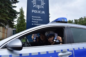 umundurowani policjant i policjantka siedzą w radiowozie i w rękach trzymają dwa odznaczenia i dwie legitymacje: „Honorowy Dawca Krwi - Zasłużony dla Zdrowia Narodu”