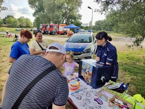 policjantka przy stoisku profilaktycznym rozmawia z dziećmi i ich opiekunami, w tle radiowóz policyjny.