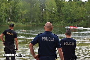 Policjanci i strażak stoją tyłem na skraju akwenu wodnego obserwując pływającą łódź.