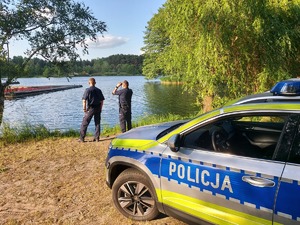 Policjanci stoją na skraju akwenu wodnego obserwując jego powierzchnię obok nich stoi radiowóz policyjny.