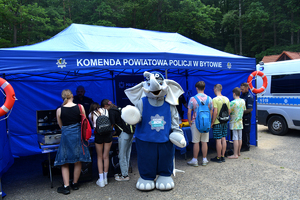 Uczestnicy festynu w stoją obok namiotu profilaktycznego, na środku maskotka policyjna.