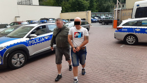 Nieumundurowany policjant prowadzi zatrzymanego oszusta zakutego w kajdanki przez parking policyjny, na którym stoją radiowozy