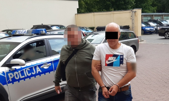 Nieumundurowany policjant prowadzi zatrzymanego oszusta zakutego w kajdanki przez parking policyjny, na którym stoją radiowozy