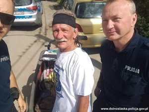 mężczyzna pozuje do zdjęcia z dwoma umundurowanymi policjantami. z tyłu widać radiowóz i cywilny samochód