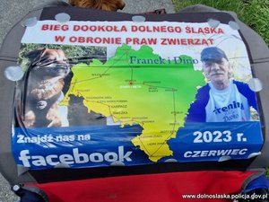 wydruk z grafiką przypięty do wózka na którym u góry widnieje czerwony napis: Bieg Dookoła Dolnego Śląska w Obronie Praw Zwierząt. Poniżej na środku znajduje się zielona mapa Dolnego Śląska, a na niej trasa biegu oraz imiona: Franek i Dino.
Po lewej stronie widoczne jest zdjęcie psa, po prawej zdjęcie Pana Franciszka. Pod spodem biały napis na niebieskim tle: Znajdź nas na facebook, 2023 r. czerwiec