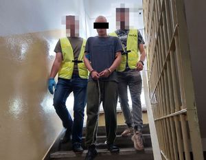 Dwaj policjanci w żółtych kamizelkach prowadzą zatrzymanego mężczyznę