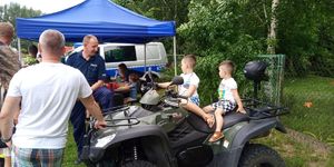Dwójka dzieci siedzi na policyjnym kładzie obok stoi ich opiekun i policjant.