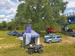 Na łące stoją namioty profilaktyczne policji w okół ustawione policyjne pojazdy służbowe i wóz strażacki.