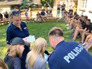 Para policjantów rozmawia z dwójką dzieci w tle grupa młodzieży siedzi na ławkach.