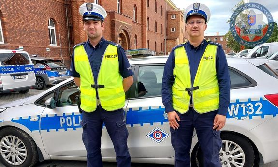 dwaj umundurowani policjanci ruchu drogowego w żółtych kamizelkach stoją przed radiowozem