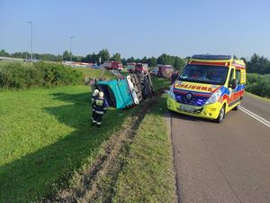 Autokar leżący w rowie. Obok na drodze stoi karetka pogotowia, a za nią wozy strażackie. Przy autobusie stoją dwaj strażacy.