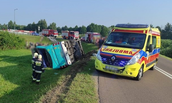 Autokar leżący w rowie. Obok na drodze stoi karetka pogotowia, a za nią wozy strażackie. Przy autobusie stoją dwaj strażacy
