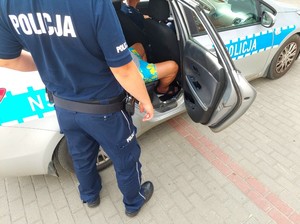 zatrzymana osoba siedzi w radiowozie na miejscu pasażera, a w otwartych drzwiach stoi policjant