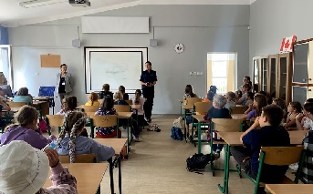Dzieci siedzące w ławkach szkolnych w klasie oraz stojąca policjantka w umundurowaniu służbowym.