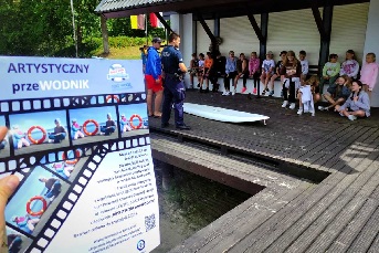 Policjant w umundurowaniu stojący przed grupą dzieci. Na pierwszym planie akcji znajduje się plakat akcji z napisem ARTYSTYCZNY PRZEWODNIK