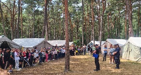 Policjantka i strażak w mundurach stojący wraz z młodzieżą w lesie, a w tle licznie rozstawione namioty.