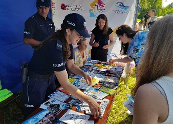 Policjantka na stanowisku policyjnym podaje dziecku kamizelkę odblaskową. Na stoliku poukładane kolorowe ulotki, broszury i malowanki. Wokół stanowiska gromadzą się dzieci