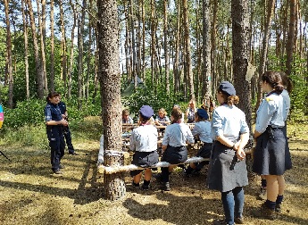 Policjantki stojące w lesie obok grupa siedzących harcerek