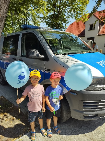 Dwaj mali chłopcy stojący z balonami w dłoni na tle radiowozu Policji.