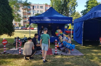 Policjant przed granatowym namiotem z napisem Komenda Powiatowa Policji w Końskich obok małych dzieci