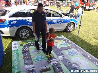 Policjantka stojąca na dużej planszy do gry z małym chłopcem. W tle radiowóz Policji.